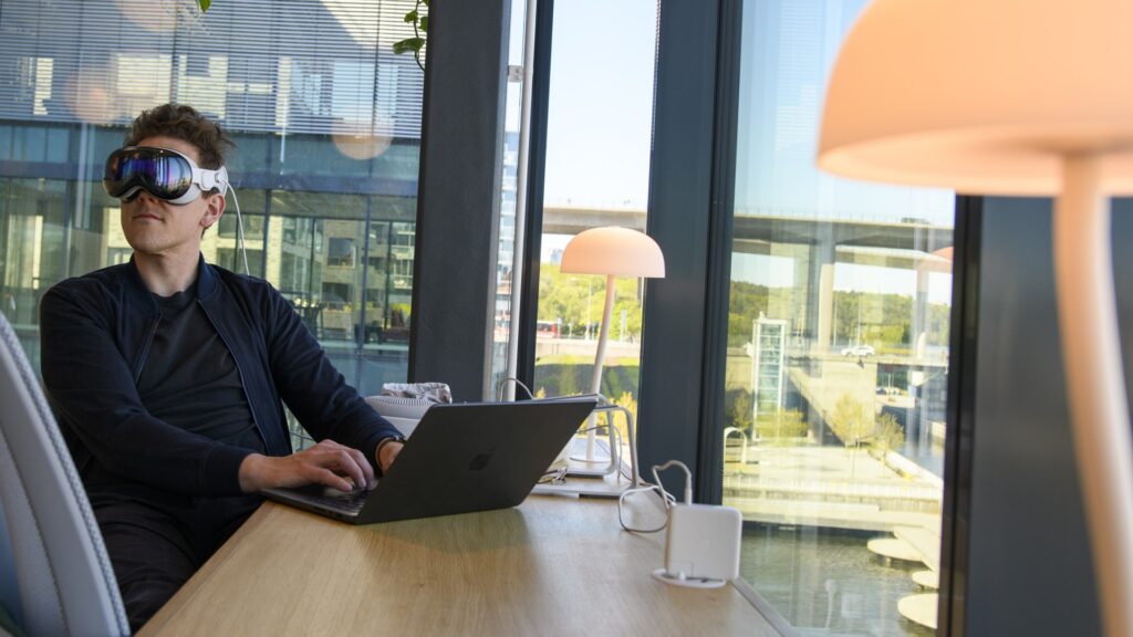 En man sitter vid ett skrivbord med en laptop på, han har VR-glasögon på sig.