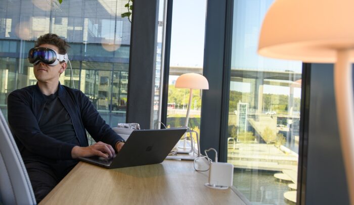 En man sitter vid ett skrivbord med en laptop på, han har VR-glasögon på sig.